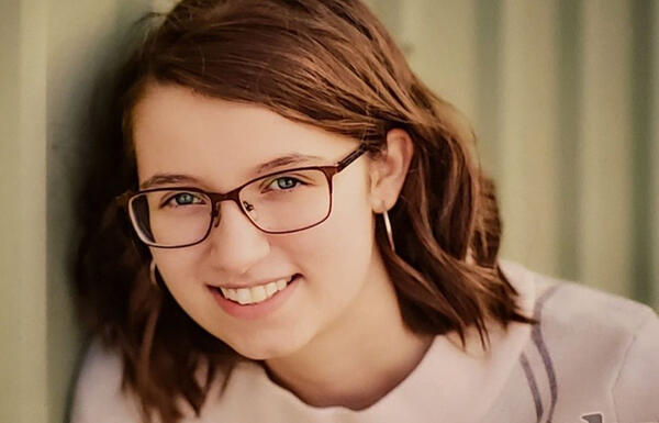 A photo of Maggie Behrendt. She is a young white woman that has wavy brown hair styled in a loose shoulder-length bob. She is also wearing large-framed glasses.